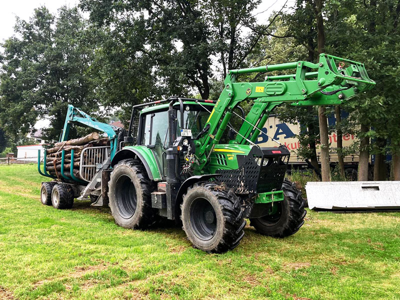 Traktor mit Holzanhänger beim Abtransport von Baumstämmen Grüner Traktor mit Anhänger und Greifarm transportiert Baumstämme über Wiese, im Hintergrund Bäume und Gebäude mit Werbung.
